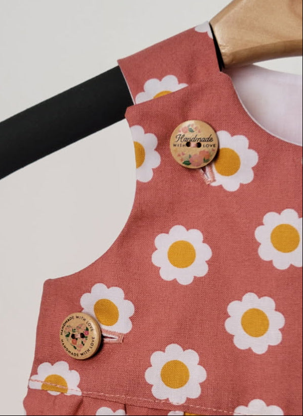 A close-up of a sleeveless children's dress or pinafore top in dusty terracotta pink, featuring an allover white daisy print with yellow centers and two decorative wooden buttons.