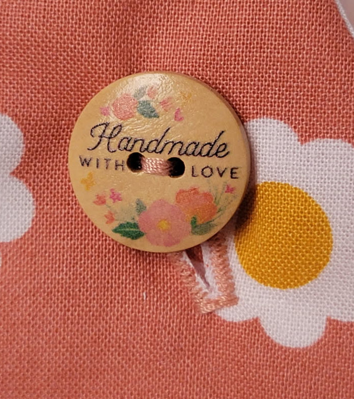 A close-up of a round wooden button with the words 'Handmade WITH LOVE' and small floral illustrations, placed on a peach fabric with a white and yellow daisy pattern.
