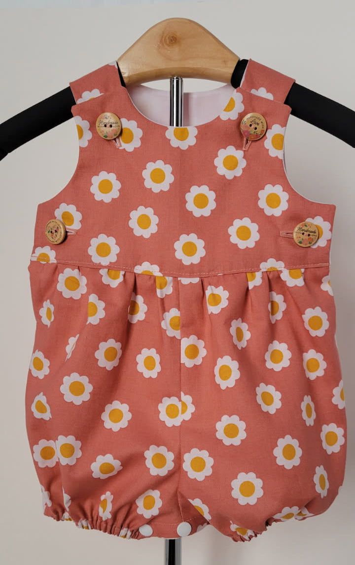 A baby romper in a dusty terracotta pink color, featuring an all-over white daisy pattern with yellow centers. It is displayed on a wooden hanger on a black hanger stand, with decorative wooden buttons on the shoulder straps.