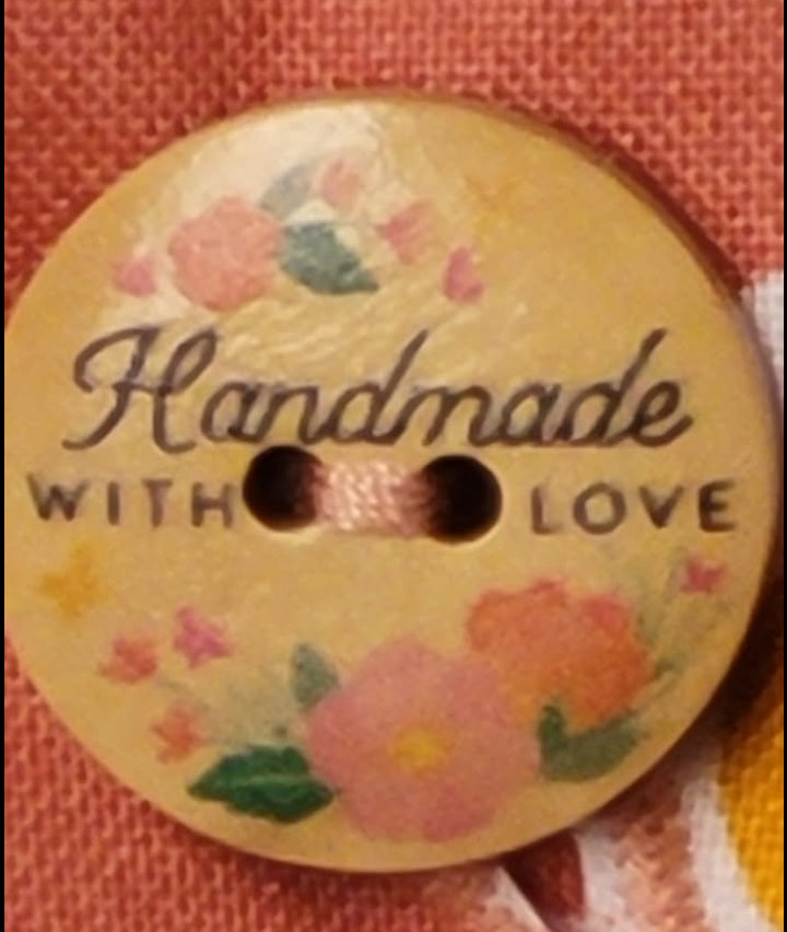 A round wooden button with two holes, featuring a hand-painted floral design in pink, green, and yellow on a light beige background. The button is printed with the words 'Handmade WITH LOVE' in black script and block lettering, and is shown resting on a textured orange fabric.