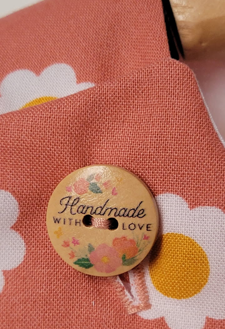 A close-up of a round wooden button with the words 'Handmade WITH LOVE' and small floral illustrations, placed on a dusty terracotta peach fabric with a white and yellow daisy pattern.