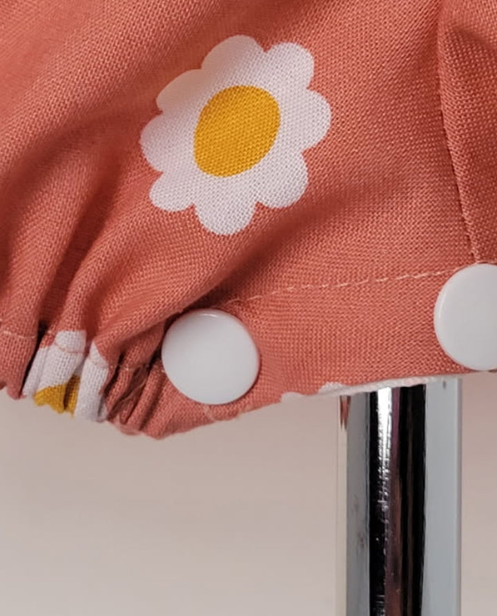 Close-up of a dusty terracotta pink fabric item with a white and yellow daisy pattern, featuring white snap buttons and a gathered edge, shown next to a silver metal stand.