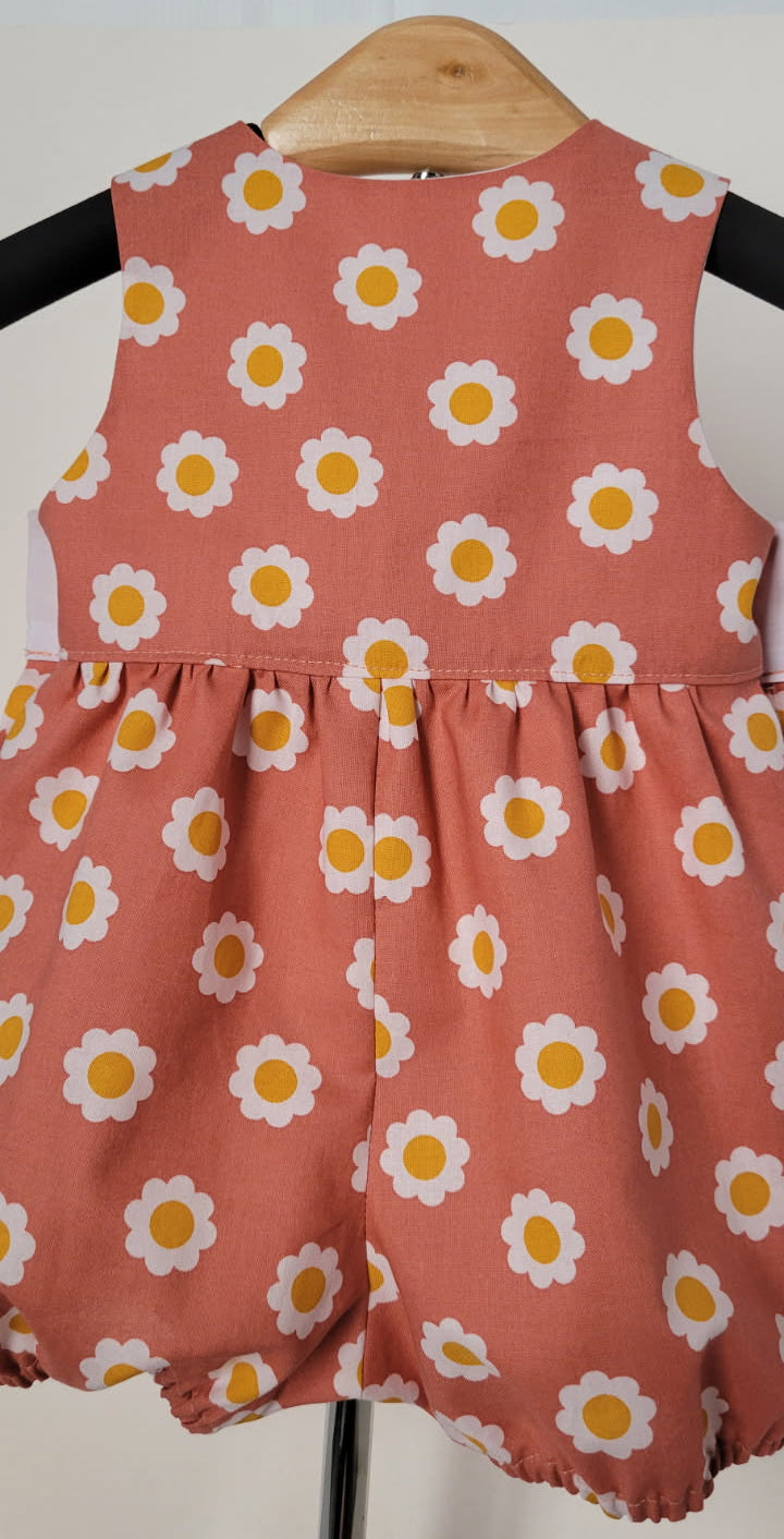 A sleeveless baby romper in a dusty terracotta pink fabric, decorated with a repeating white daisy pattern with yellow centers, hanging on a wooden hanger.
