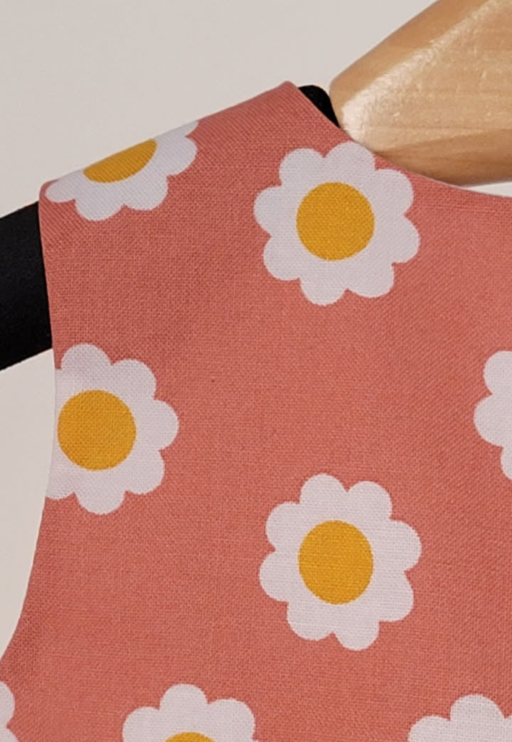 A close-up of a dusty terracotta pink fabric product, likely a sleeve or strap, featuring a repeating pattern of white daisy flowers with yellow centers, shown on a wooden hanger.