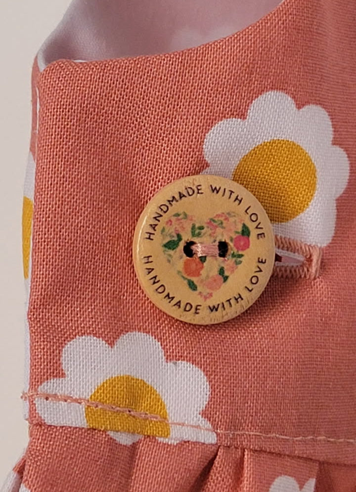Close-up of a dusty terracotta pink fabric garment with a daisy print and a round wooden button that reads 'HANDMADE WITH LOVE' and features a small floral design.