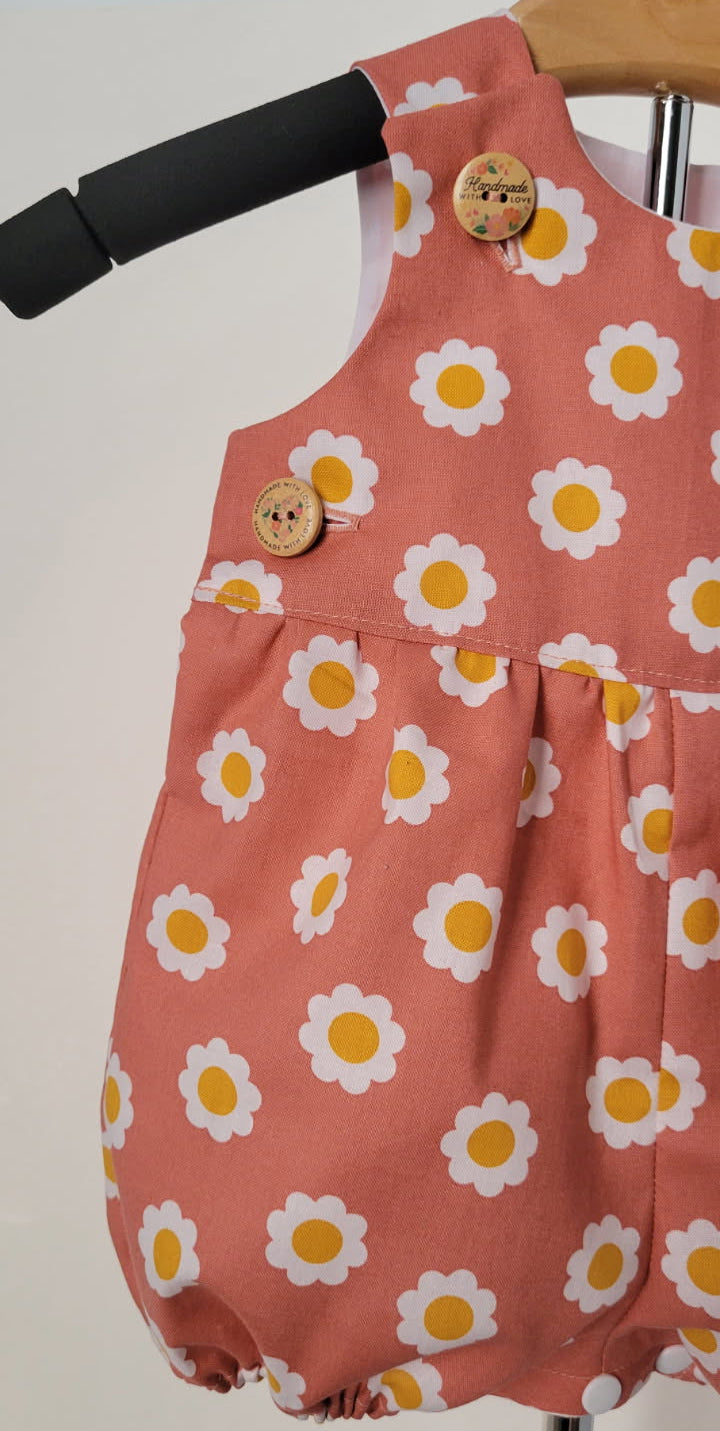 A baby romper in dusty terracotta pink with a white and yellow daisy print, featuring two decorative wooden buttons, hanging on a wooden hanger.