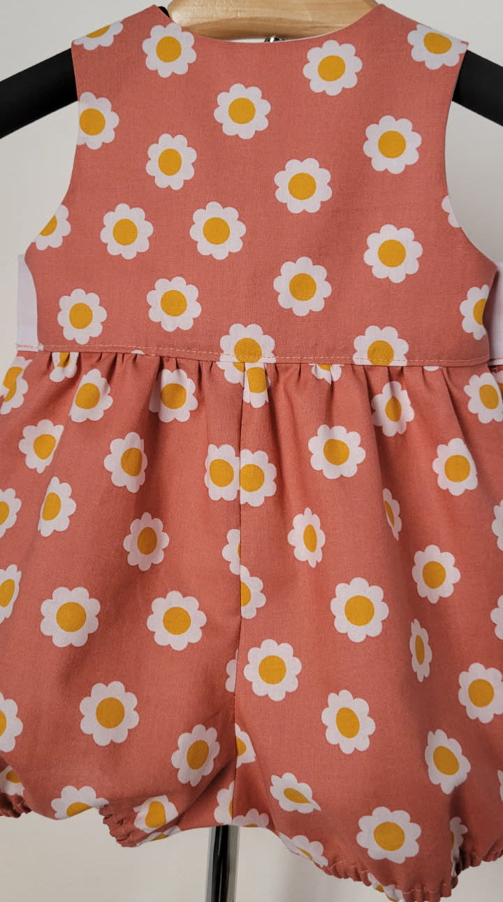 A sleeveless baby romper in a dusty terracotta peach fabric, decorated with a repeating white daisy pattern with yellow centers, hanging on a wooden hanger.