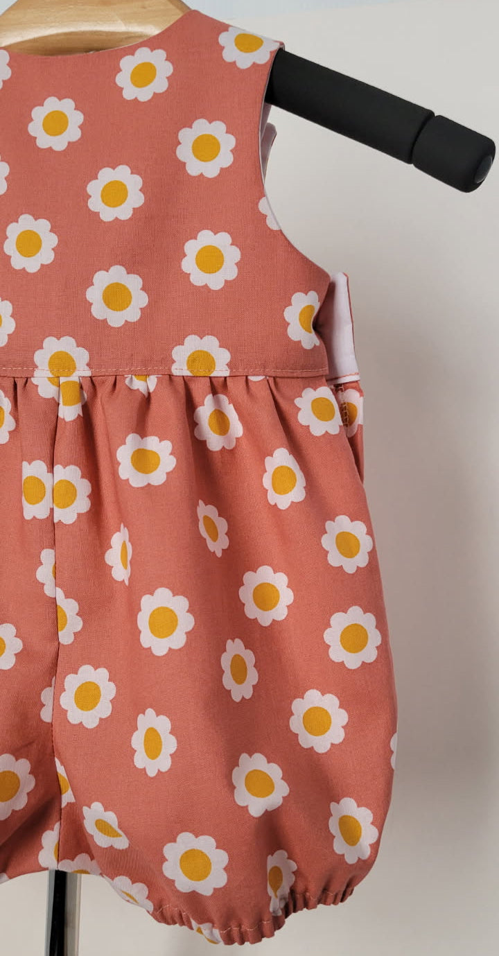 A sleeveless children's romper in a dusty terracota peach color with a white and yellow daisy print, displayed on a black hanger against a plain light background.